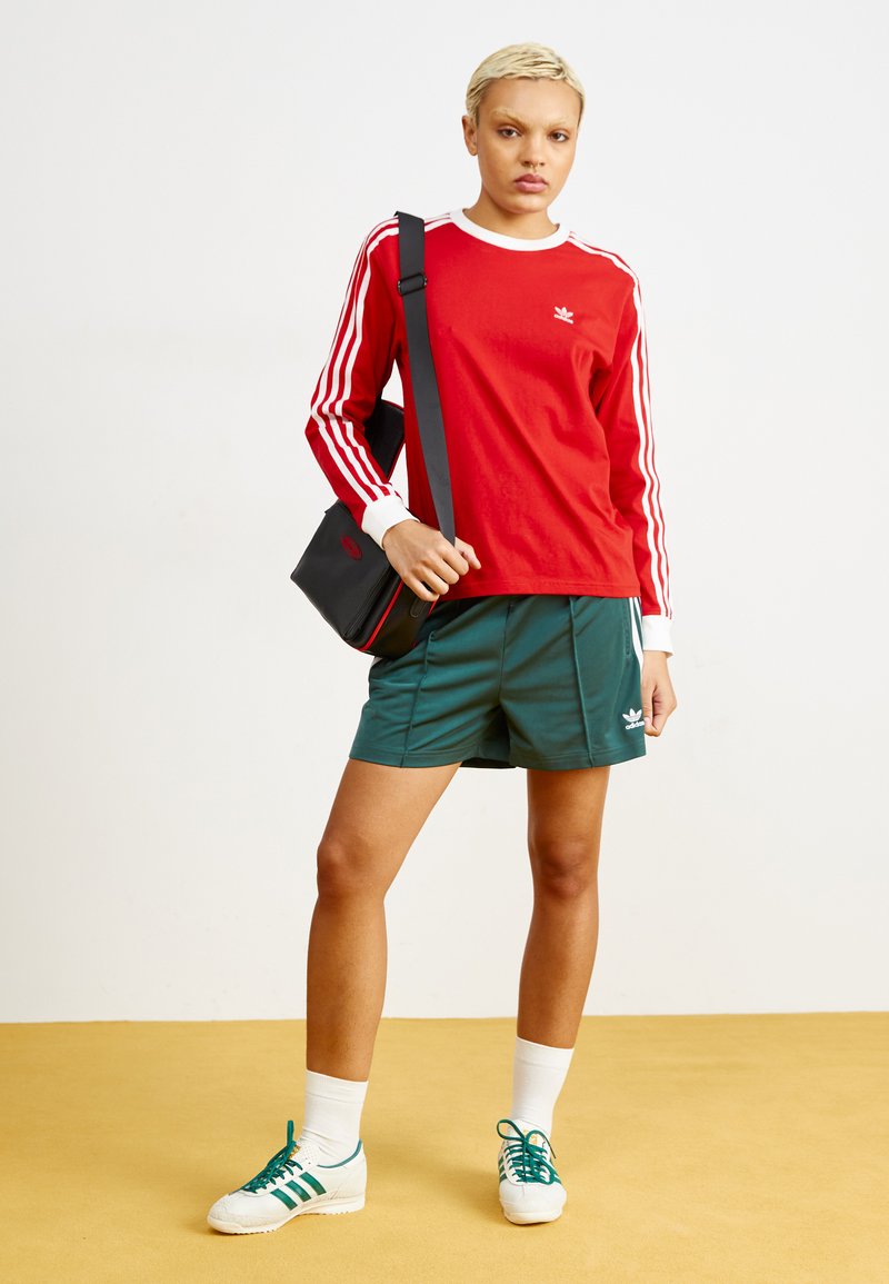 Chemise rouge à manches longues avec des rayures blanches, shorts verts, chaussettes blanches et chaussures avec des accents verts. Sac noir avec des détails rouges. Fond jaune.