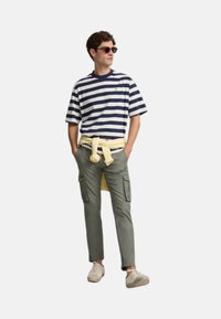 Camiseta a rayas azul marino y blanca, pantalones cargo caqui con bolsillos, corbata amarilla en la cintura y zapatillas ligeras. Las gafas de sol redondas completan el look.