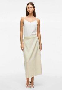 Camisole blanche à fines bretelles, associée à une longue jupe de couleur beige clair. Tissu lisse avec un léger éclat, design minimaliste et talons avec bride au niveau de la cheville.
