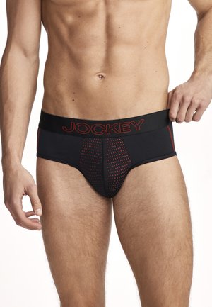 Männlicher Oberkörper mit schwarzen Slips mit rotem Punktmuster und „JOCKEY“-Logo am Bund, rechte Hand stellt den Bund ein.