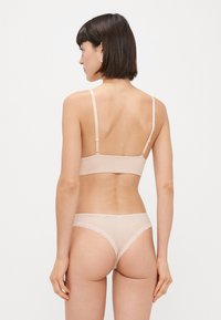 Naha bralette õhukeste rihmadega ja siledast materjalist, koos ühte suguset thongiga, millel on pitsäär. Lihtne, minimalistlik disain.