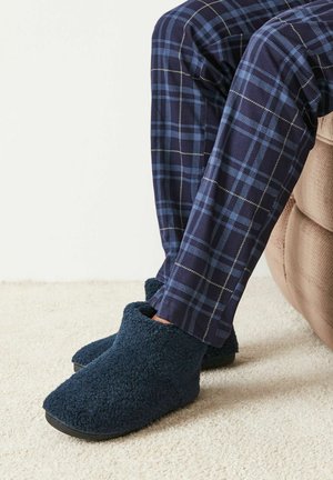 Navy fuzzy slippers with a soft texture and rounded toe, paired with plaid pants in navy and blue tones, resting on a light carpet.