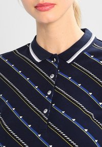 Polo shirt blu navy con colletto bianco, caratterizzato da strisce diagonali multicolori e piccoli motivi a cuore. I bottoni si trovano sul colletto.