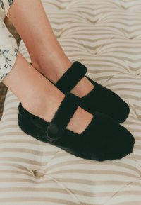 Czarne, puszyste kapcie z płaską podeszwą i zaokrąglonym czubkiem, z paskiem z przodu i detalem w postaci guzika, umieszczone na teksturowanej, prążkowanej powierzchni.