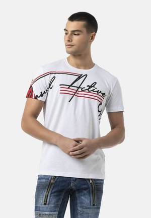 Junger Mann trägt ein weißes T-Shirt mit schwarzem und rotem "Active" Text und blaue Jeans mit vertikalen Reißverschluss-Details, steht mit verschränkten Händen.