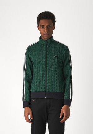 Chaqueta verde con cremallera y patrón de diamantes, con franjas blancas a lo largo de las mangas y un dobladillo y puños de canalé negros. Logo de Lacoste presente.