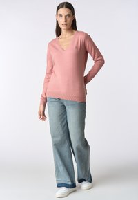 Maglione rosa a scollo a V con maniche lunghe, orlo a coste e piccolo logo. Abbinato a jeans larghi blu chiaro e sneaker bianche.