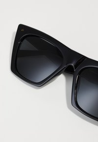 Mustat ylisuurent aurinkolasit, joissa on kulmikas muotoilu, tummat linssit ja kiiltävä pinta. Minimalistinen varustus, jossa on pieni logo toisessa sankassa.