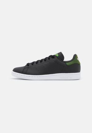 Black low-top sneaker with white sole, black laces, and green-yellow striped heel tab and tongue detailing.