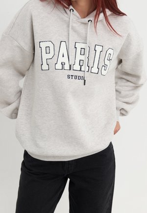 Kobieta ubrana w jasnoszarą bluzę z kapturem z dużym napisem "PARIS" oraz czarne spodnie na jednolitym tle.