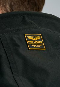 Tessuto nero con un dettaglio di cucitura triangolare e un'etichetta giallo e nera che recita "PME LEGEND" con ali e "TESTATO A ALTEZZA SOFFICE."