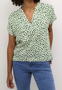 Woman wearing short-sleeve white blouse with green and black spots, paired with blue jeans, standing against a plain background.