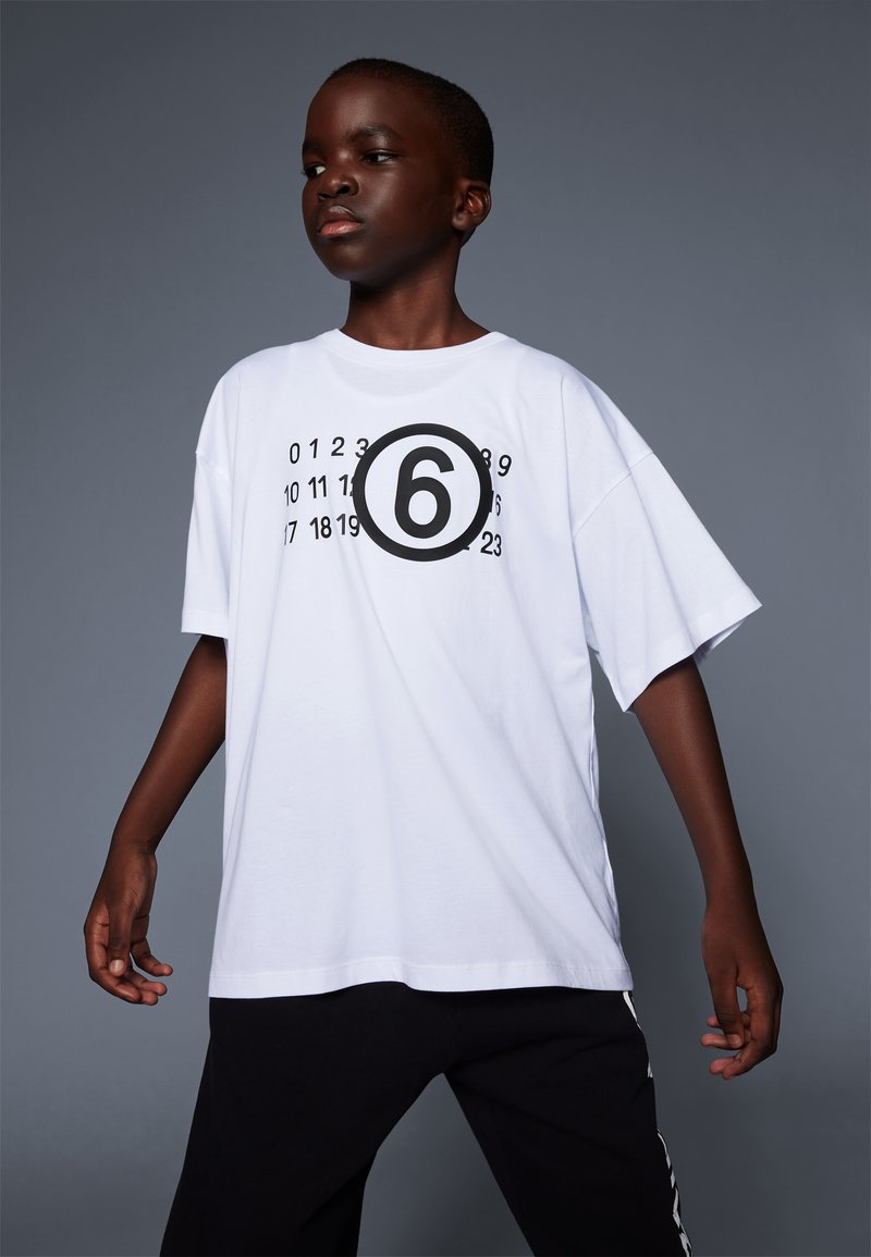 T-shirt bianco in cotone con vestibilità oversize, caratterizzato da un grande cerchio nero con il numero "6" al centro, circondato da piccoli numeri.