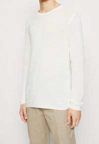 Pull en maille blanche avec un motif texturé, col rond, et poignets ajustés, associé à un pantalon beige.