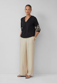 Zwarte blouse met een V-hals en driekwart mouwen, voorzien van gouden geborduurde accenten. Brede, lichtbeige pantalon met een gestructureerde afwerking.