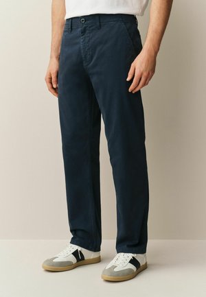 Mann trägt dunkelblaue gerade geschnittene Hose und weiße Sneaker mit schwarzen und grauen Akzenten vor einem einfarbigen beigen Hintergrund.