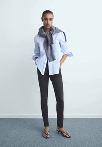 Mujer de pie en interiores, vistiendo una camisa de botones azul claro, suéter gris plegado sobre los hombros, pantalones negros ajustados y tacones negros de tiras.