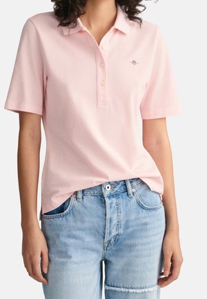 Woman wearing light pink short-sleeve polo shirt tucked into high-waist light blue denim jeans with a patch detail on the thigh.