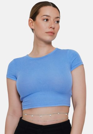 Giovane donna con i capelli legati indossa un crop top blu e catene d'argento a strati in vita, guarda verso destra su uno sfondo semplice.