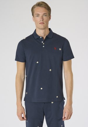 Uomo che indossa una polo blu navy e pantaloni con piccoli motivi di margherite bianche, in piedi davanti a uno sfondo chiaro e uniforme.
