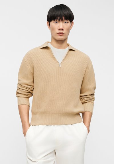 Μπεζ πλεκτό πουλόβερ με γιακά με μισό φερμουάρ και ριμπ μανσέτες, με υφασμάτινο σχέδιο, φορεμένο πάνω από ένα λευκό πουκάμισο και συνδυασμένο με ανοιχτόχρωμο παντελόνι.