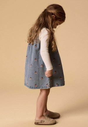 Jeune fille en robe bleue à motifs de cerises regardant vers le bas, portant un chemisier blanc à manches longues et des chaussures beige à enfiler sur un fond beige.