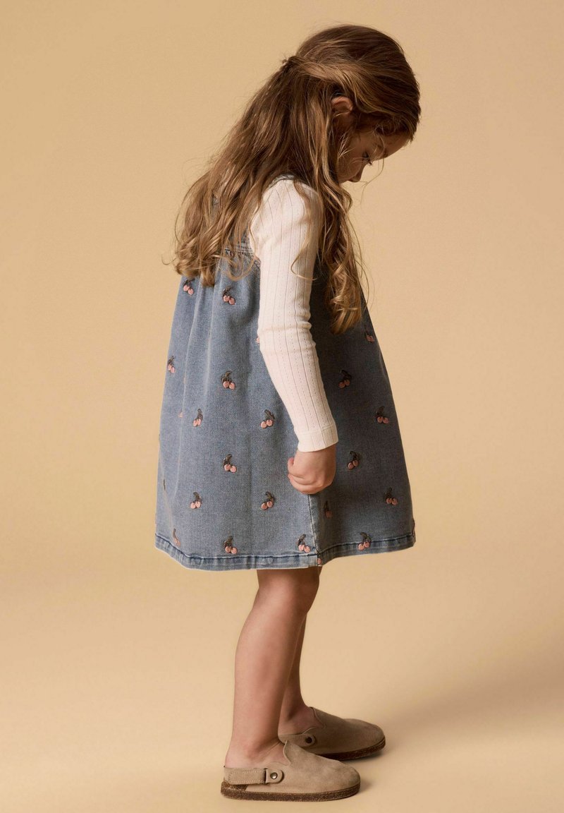 Nuori tyttö sinisessä mekossa, jossa on kirsikkakuviot, katsoo alas. Hänellä on valkoinen pitkähihaisen paita ja beiget liukuselkätossut vaaleanruskealla taustalla.