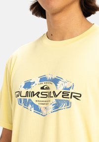 Camiseta amarilla de manga corta con un gráfico floral azul y blanco que incluye el logotipo de "Quiksilver" y un diseño hexagonal. Material de algodón suave.
