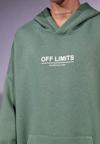 Grønn hettegenser laget av myk bomull, med en frontlomme og hvit tekst "OFF LIMITS DANGEROUS AREA" på brystet.