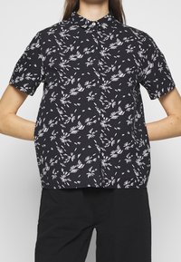 Camisa negra de manga corta con un patrón floral blanco, con un cierre de botones al frente y un diseño de cuello clásico. Tejido ligero.