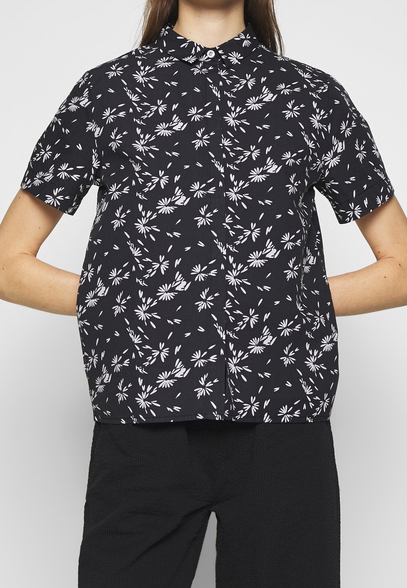Camisa negra de manga corta con un patrón floral blanco, con un cierre de botones al frente y un diseño de cuello clásico. Tejido ligero.