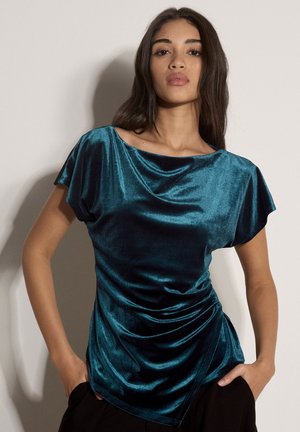 Femme portant un top court en velours brillant couleur bleu sarcelle avec un léger drapé, debout les mains dans les poches d’un pantalon noir devant un fond uni.