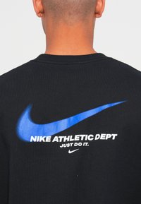 Fekete pamut póló nagy kék Nike logóval és fehér felirattal: "NIKE ATHLETIC DEPT JUST DO IT." a hátsó részen.