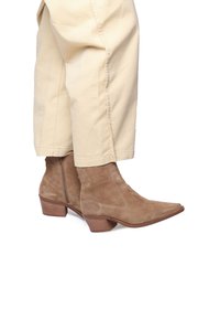 Botas de ante marrón hasta el tobillo con punta afilada y tacón bajo apilado, que presentan un cierre lateral con cremallera y una textura suave, combinadas con pantalones de color claro.