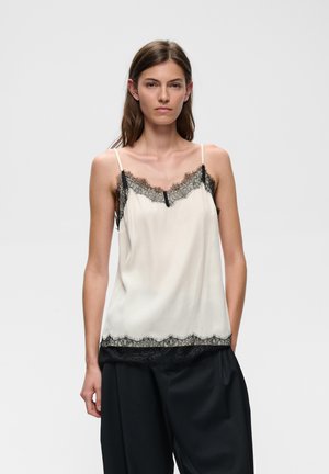 Femme aux cheveux bruns et raides porte un haut en satin blanc sans manches avec une bordure en dentelle noire et un pantalon noir ample, debout devant un fond uni.