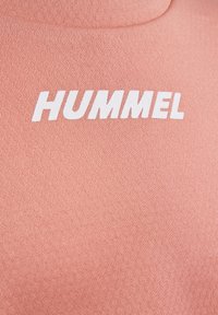 Close-up van roze gestructureerde stof met het witte, vette "HUMMEL"-logo gedrukt op het borstgebied van een kledingstuk.