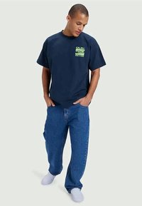 Navy blue cotton t-shirt with a neon green graphic logo on the chest, paired with loose blue denim jeans and white sneakers.