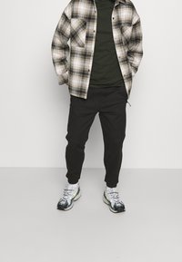 Plédová flanelová košile v béžové a černé barvě se dvěma náprsními kapsami, doplněná tmavě zeleným tričkem a černými zúženými tepláky. Obsahuje sportovní tenisky.