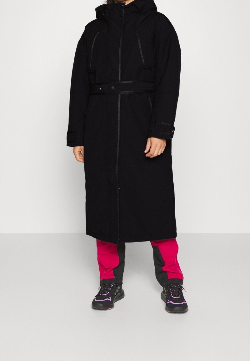 Lång svart kappa med huva, med dragkedja fram och bälte. Bärs över rosa och svarta byxor, kombinerat med träningsskor.