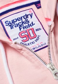 Sudadera de color rosa claro con una cremallera blanca y una etiqueta azul, roja y blanca que dice "Superdry Track & Field" y "Athletic Dept.".