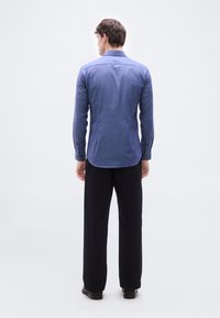 Άνδρας που στέκεται με την πλάτη στραμμένη, φοράει μπλε μακρυμάνικο πουκάμισο, μαύρο παντελόνι και μαύρα παπούτσια, με φόντο λευκό και απλό.