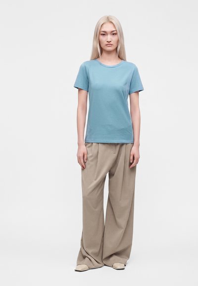 Femme aux longs cheveux blonds portant une chemise bleue à manches courtes et un pantalon large beige, debout devant un fond clair uni.