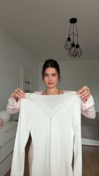 Långärmad vit topp med spetsdetalj vid halsringningen, med en mjuk textur och avslappnad passform, hålls uppe mot en neutral bakgrund.