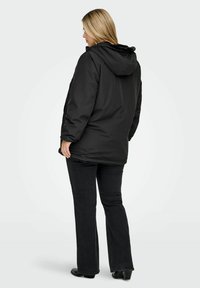 Sieviete, which wears a black hooded winter jacket and dark jeans, standing in a three-quarter back view against a plain light background.