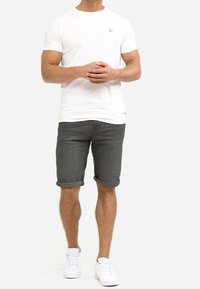 Camiseta blanca de manga corta con un pequeño logo, combinada con pantalones cortos de mezclilla gris oscuro arremangados y zapatillas blancas. Conjunto simple y casual.