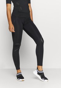 Osoba ubrana w czarne legginsy sportowe i czarne buty wsuwane z białą podeszwą, stojąca na tle jasnego, jednolitego tła.