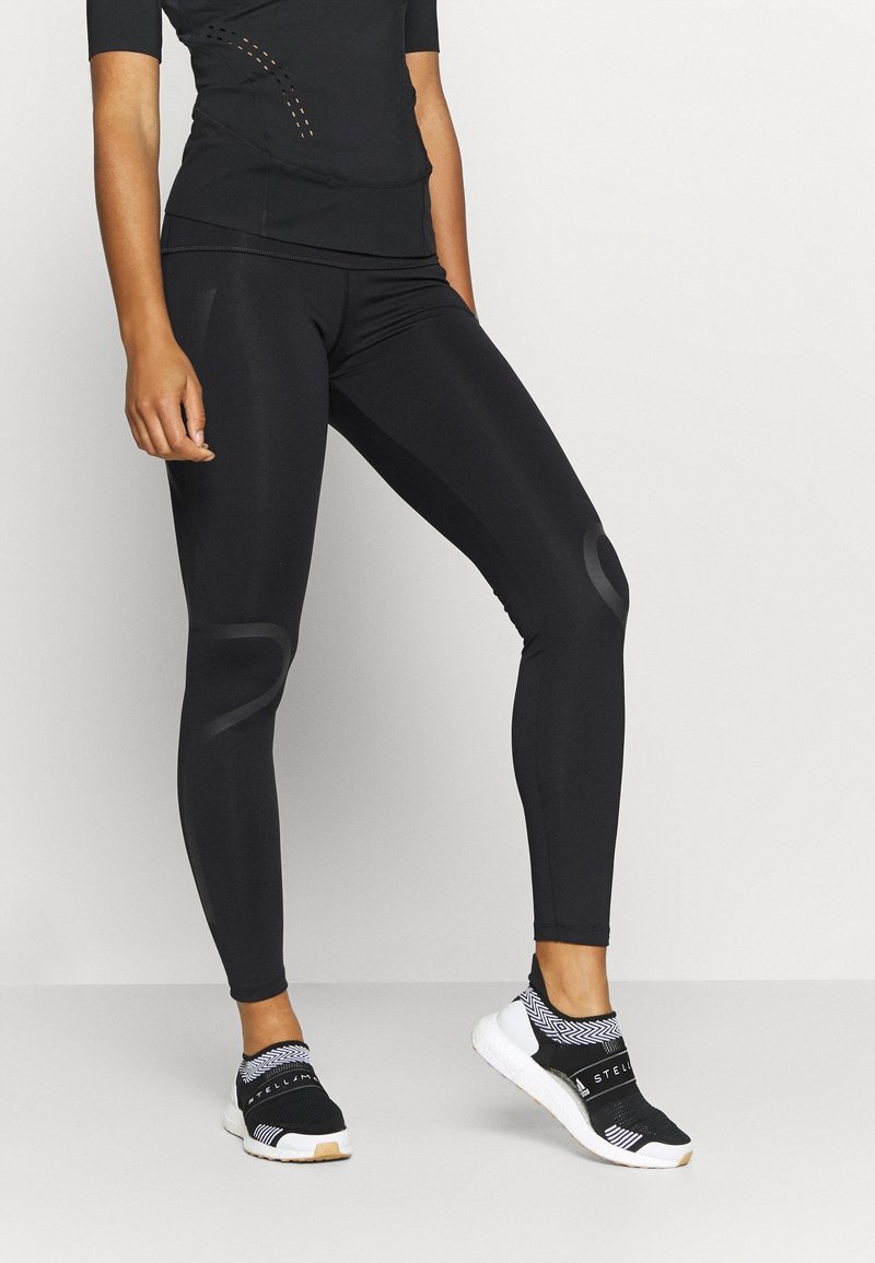 Osoba ubrana w czarne legginsy sportowe i czarne buty wsuwane z białą podeszwą, stojąca na tle jasnego, jednolitego tła.