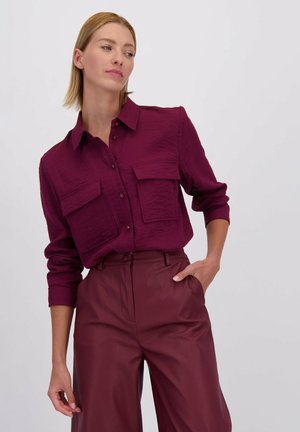 Bordeaux overhemd met een gestructureerde afwerking, voorzien van twee voorzakken en lange mouwen; gecombineerd met hoog opgesneden bordeaux faux leren broek.