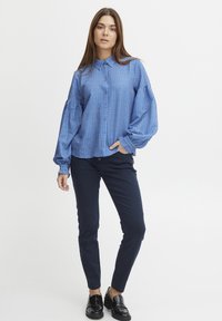 Camicia blu a fantasia con maniche a sbuffo, abbinata a pantaloni navy attillati e scarpe nere slip-on. Tessuto semplice e liscio.