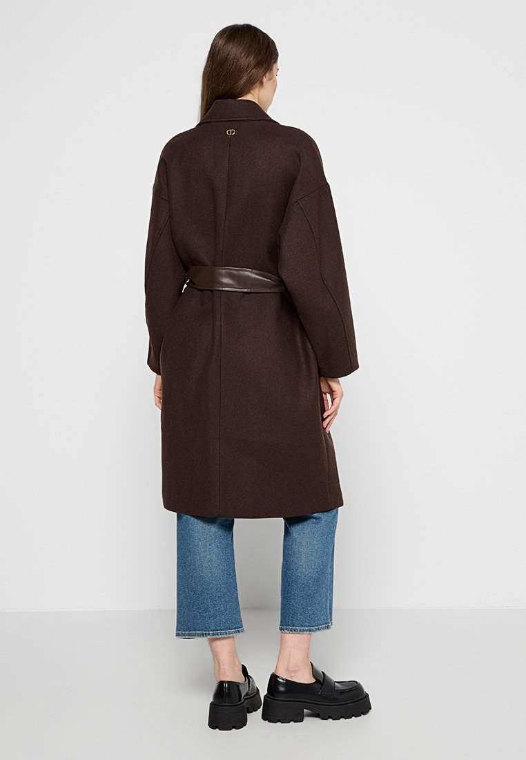 Brauner Wollmantel mit einem taillierten Gürtel, überschnittenen Schultern und einem Rückendetail mit Nadel. Kombiniert mit hellblauen gerade geschnittenen Jeans und schwarzen Plateauschuhen.